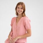 Mujer sonriente con blusa rosa de manga corta y botones, combinada con un bolso del mismo color, fondo blanco. Ideal para moda femenina y estilo casual.