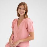 Mujer sonriente con blusa rosa de manga corta y botones, combinada con un bolso del mismo color, fondo blanco. Ideal para moda femenina y estilo casual.