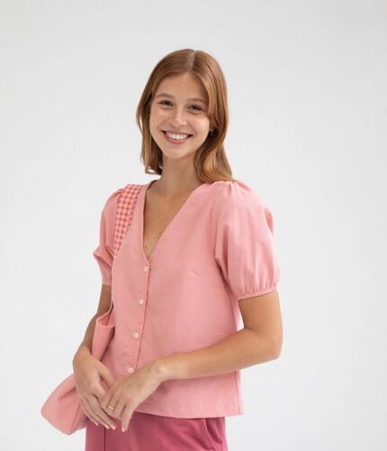 Mujer sonriente con blusa rosa de manga corta y botones, combinada con un bolso del mismo color, fondo blanco. Ideal para moda femenina y estilo casual.