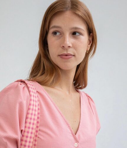 Mujer con blusa de cuadros rosas y tirantes a rayas, mirando hacia un lado, fondo neutro.