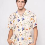 Hombre joven posando con una camisa de estampado abstracto en tonos beige, azul, naranja y verde, luciendo un estilo moderno y casual.