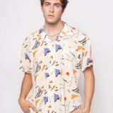 Hombre joven posando con una camisa de estampado abstracto en tonos beige, azul, naranja y verde, luciendo un estilo moderno y casual.