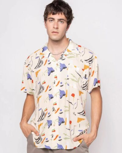 Hombre joven posando con una camisa de estampado abstracto en tonos beige, azul, naranja y verde, luciendo un estilo moderno y casual.