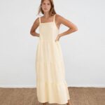 Mujer joven posando con un vestido largo amarillo de tirantes en un fondo blanco, destacando un estilo fresco y veraniego. Ideal para ocasiones casuales.