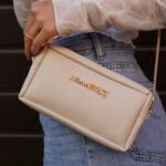 Mujer sosteniendo un bolso de mano beige con cadena dorada, luciendo una blusa blanca y jeans, en un entorno urbano.