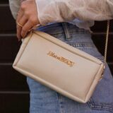 Mujer sosteniendo un bolso de mano beige con cadena dorada, luciendo una blusa blanca y jeans, en un entorno urbano.