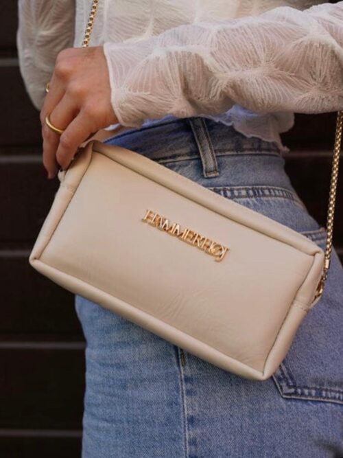 Mujer sosteniendo un bolso de mano beige con cadena dorada, luciendo una blusa blanca y jeans, en un entorno urbano.