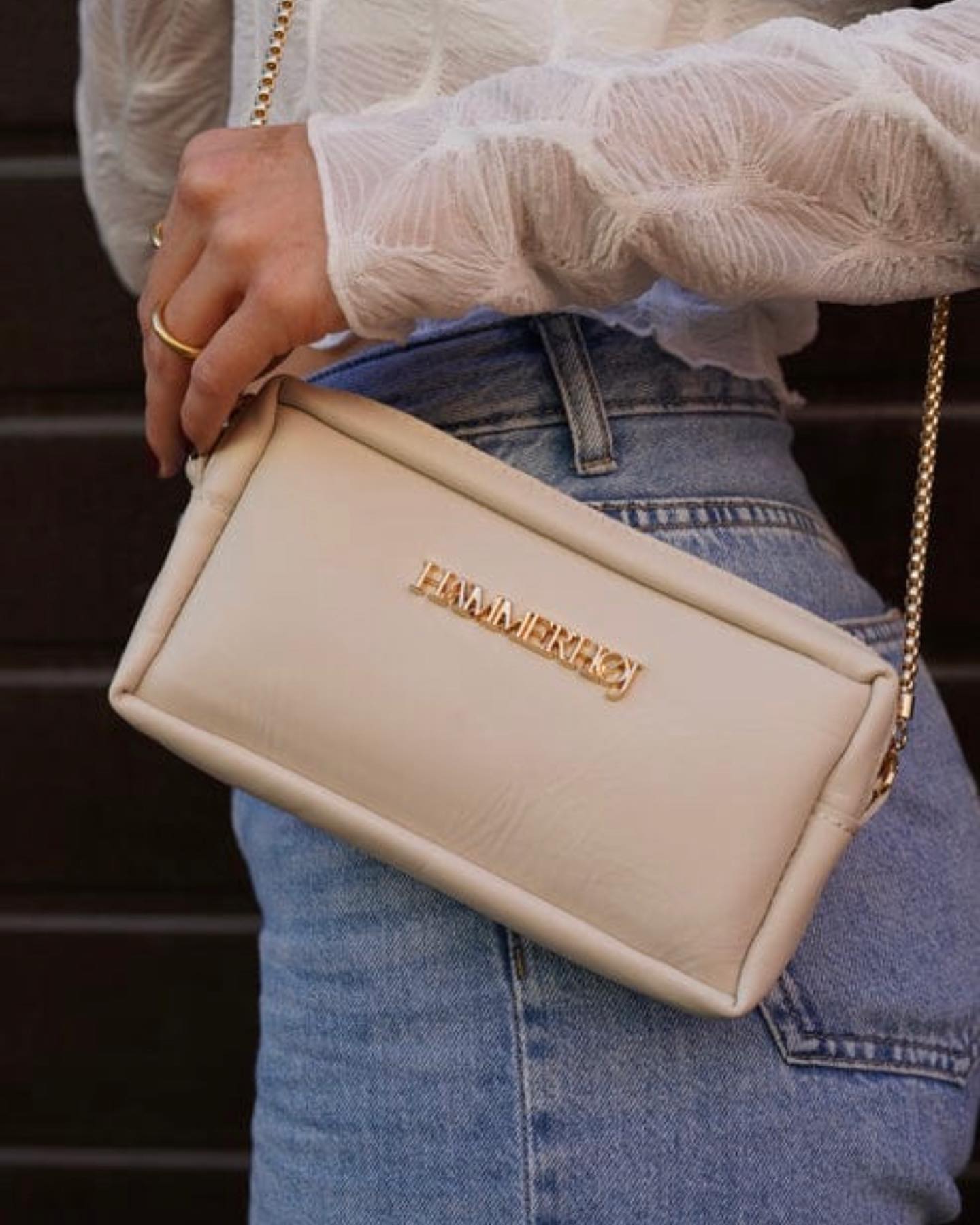 453315443_18362567626098092_3810997860928139445_n Mujer sosteniendo un bolso de mano beige con cadena dorada, luciendo una blusa blanca y jeans, en un entorno urbano.