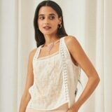 Mujer posando con blusa sin mangas de color blanco con bordados delicados y detalles de encaje, complementada con un collar de piedras rosas, en un entorno con fondo claro.