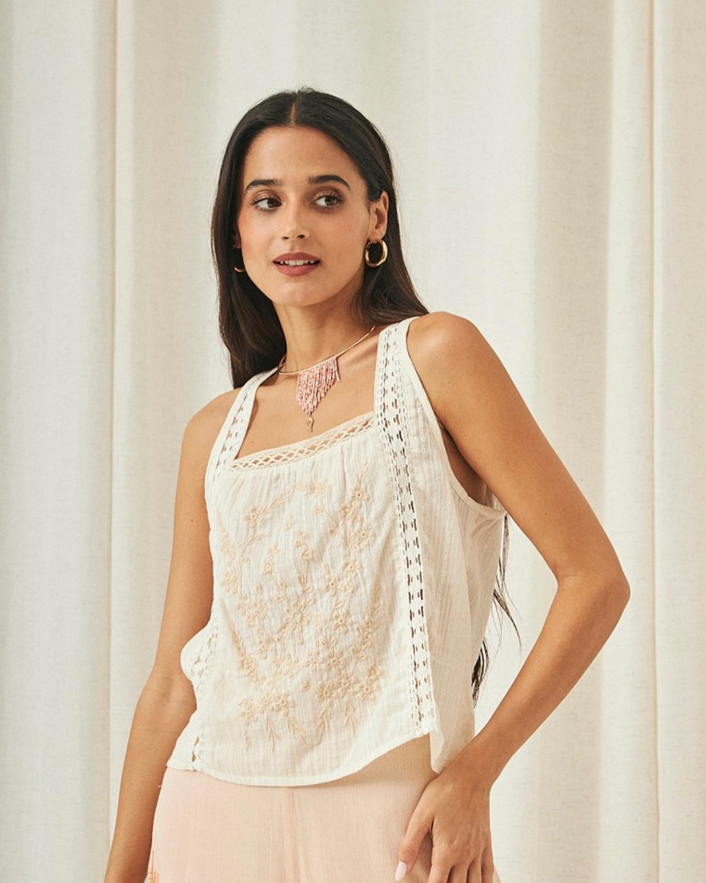 453617126_18362999827098092_1976653740058244197_n Mujer posando con blusa sin mangas de color blanco con bordados delicados y detalles de encaje, complementada con un collar de piedras rosas, en un entorno con fondo claro.