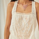 Mujer luciendo una blusa sin mangas de color blanco con bordados florales en tonos beige, combinada con un collar de cuentas en tonos rosados y dorados. Fondo neutro que resalta la prenda.