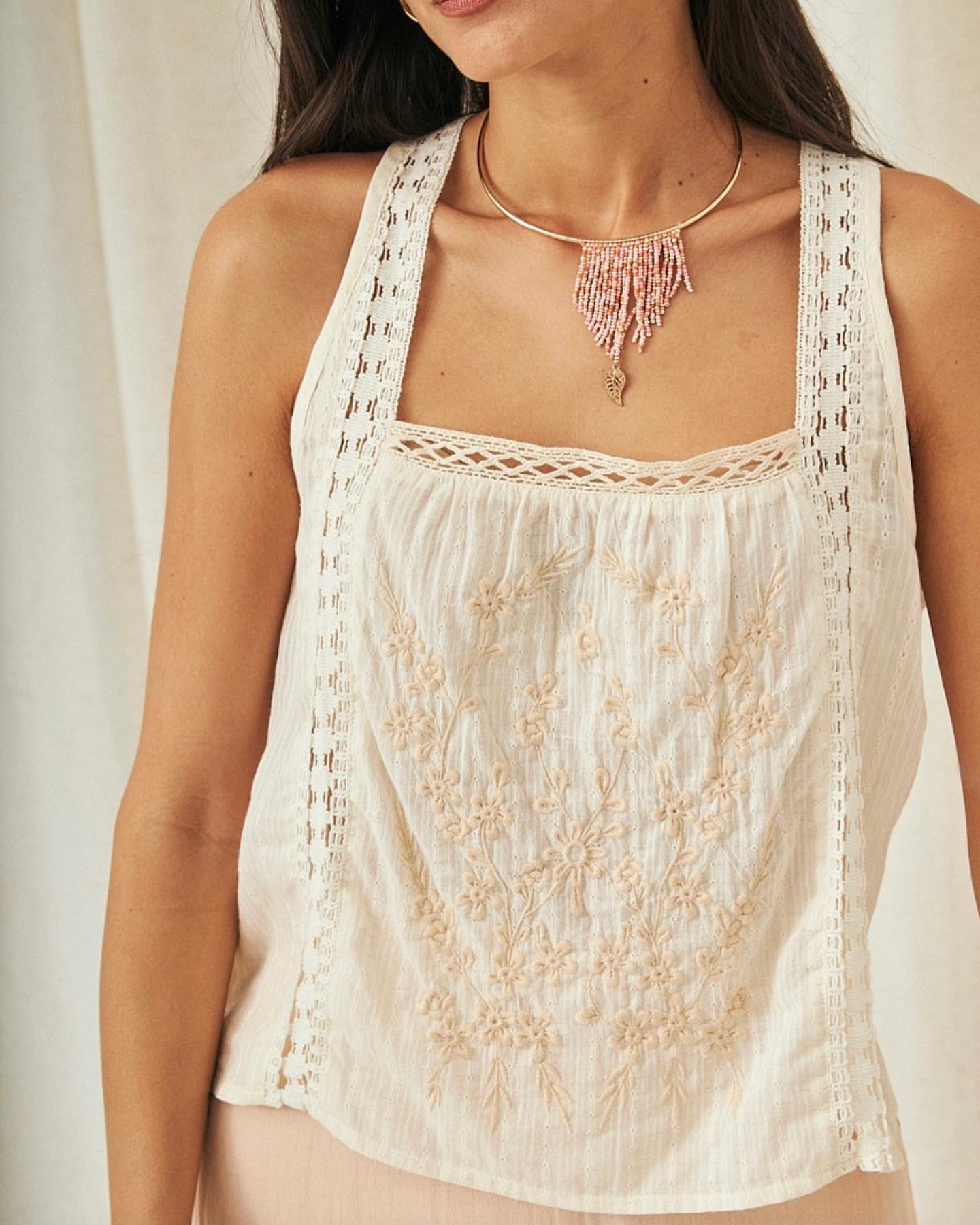 453723560_18362999839098092_4706540130008115647_n Mujer luciendo una blusa sin mangas de color blanco con bordados florales en tonos beige, combinada con un collar de cuentas en tonos rosados y dorados. Fondo neutro que resalta la prenda.